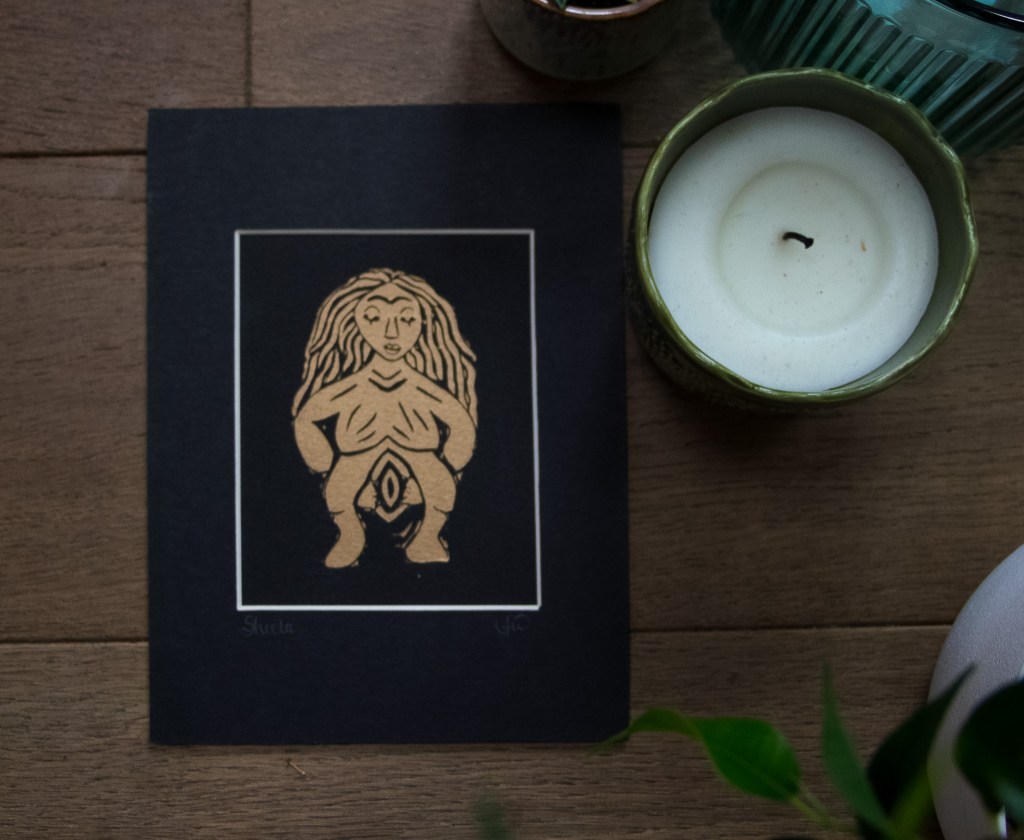 A linocut print of Sheela na Gig, depicting a goddess figure with exaggerated features, set against a black background, surrounded by a candle and plants.