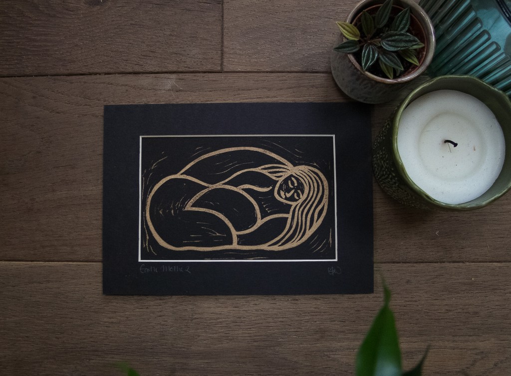 A linocut print depicting Gaia, the personification of Earth, with flowing hair and a nurturing posture, surrounded by decorative elements like a candle and small plants on a wooden surface.