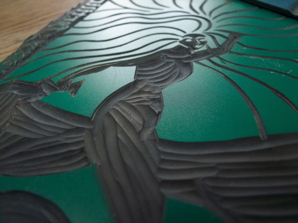 Close-up of a linocut print depicting the Goddess Brigid, characterized by flowing lines representing fire and inspiration, set against a green background.