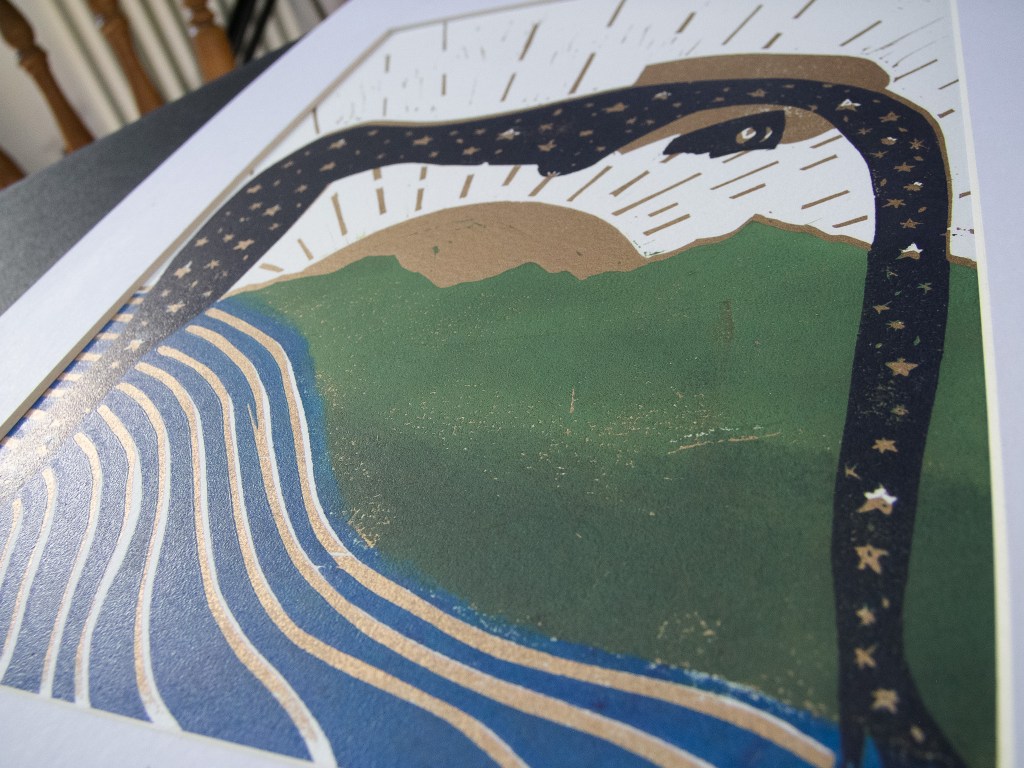 An artistic linocut depicting a stylized serpent with a starry body curving over green hills and wavy blue lines, surrounded by rays of light.