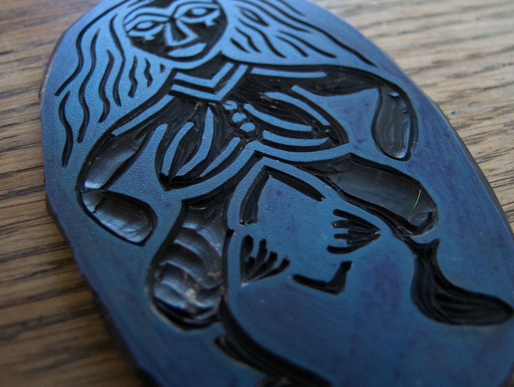 Close-up image of a carved linocut of Sheela na Gig, depicting a female figure with long hair and a prominent vulva, symbolizing fertility and femininity. The carving has intricate details, showcasing a dramatic pose that reflects her duality as a goddess of life and death.