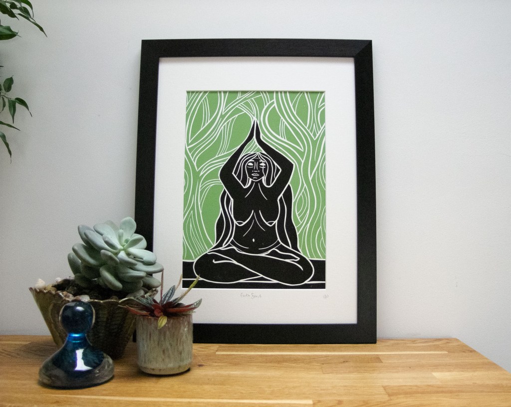 A framed linocut artwork featuring a woman in a meditative pose, surrounded by abstract green patterns resembling nature, with succulents and decorative items on a wooden surface.