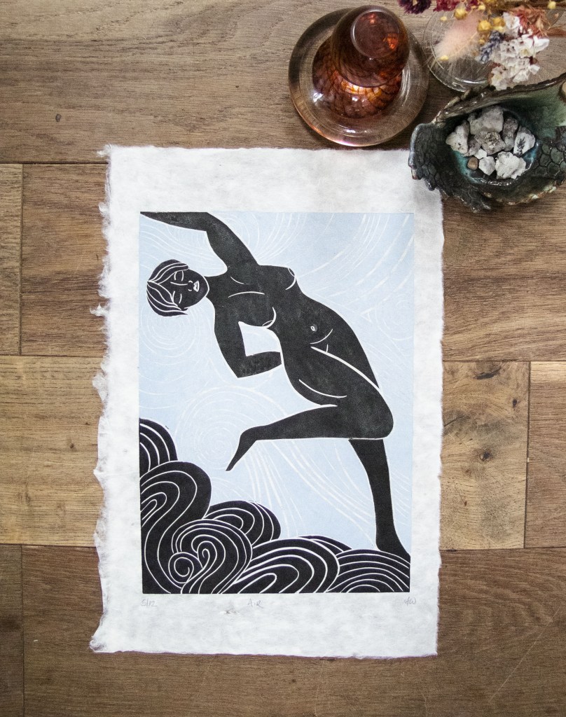 A linocut print depicting a stylized figure of a woman in a dynamic pose against a light blue background, surrounded by black swirling waves. The artwork is presented on textured paper and sits on a wooden surface with decorative items like a glass and a small container.