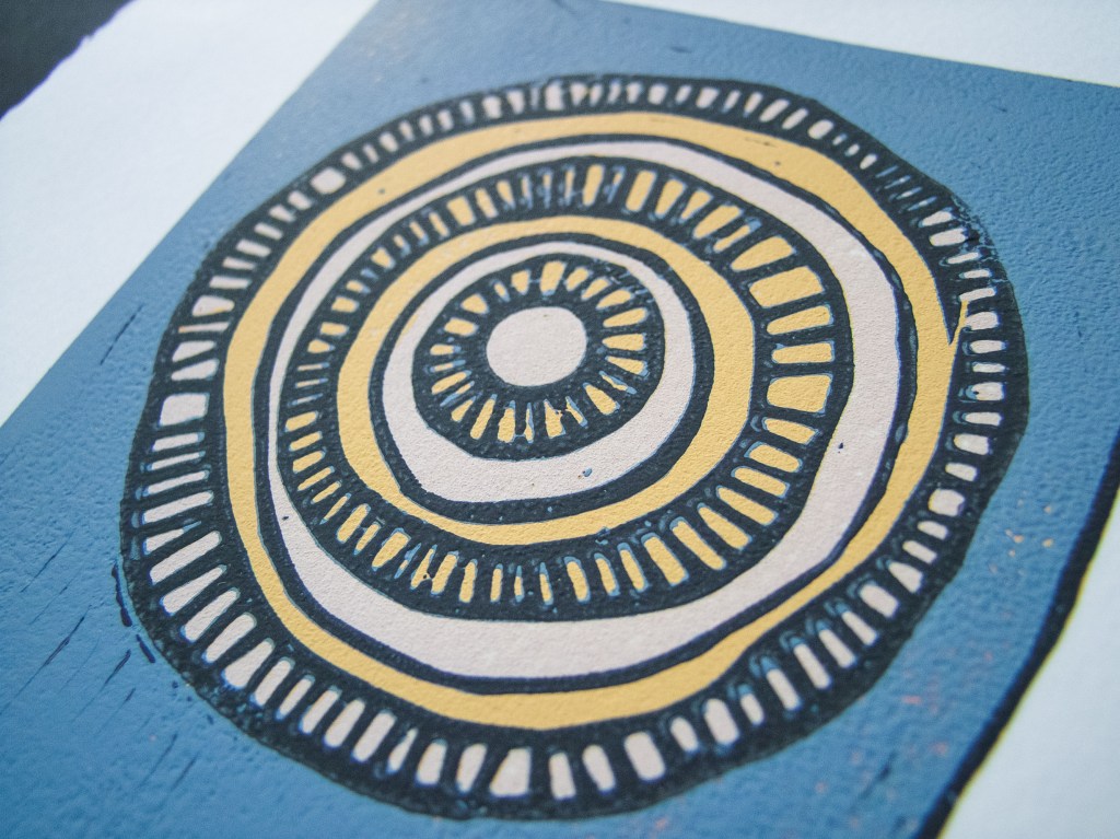 A close-up of a circular linocut print featuring concentric circles in various colors, predominantly blue and beige, with textured details.