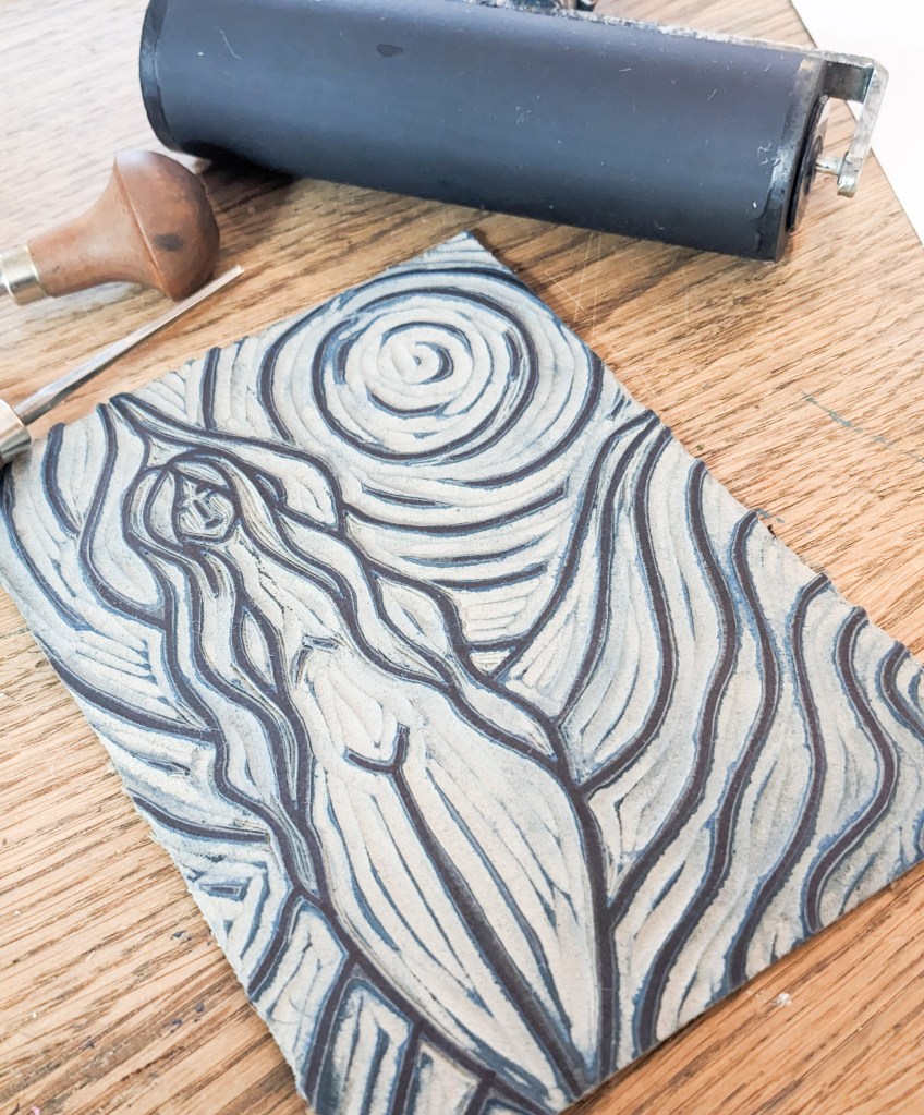 A hand-printed linocut of Goddess Achelois, featuring flowing lines and a swirling background, alongside a linocut tool and roller on a wooden surface.
