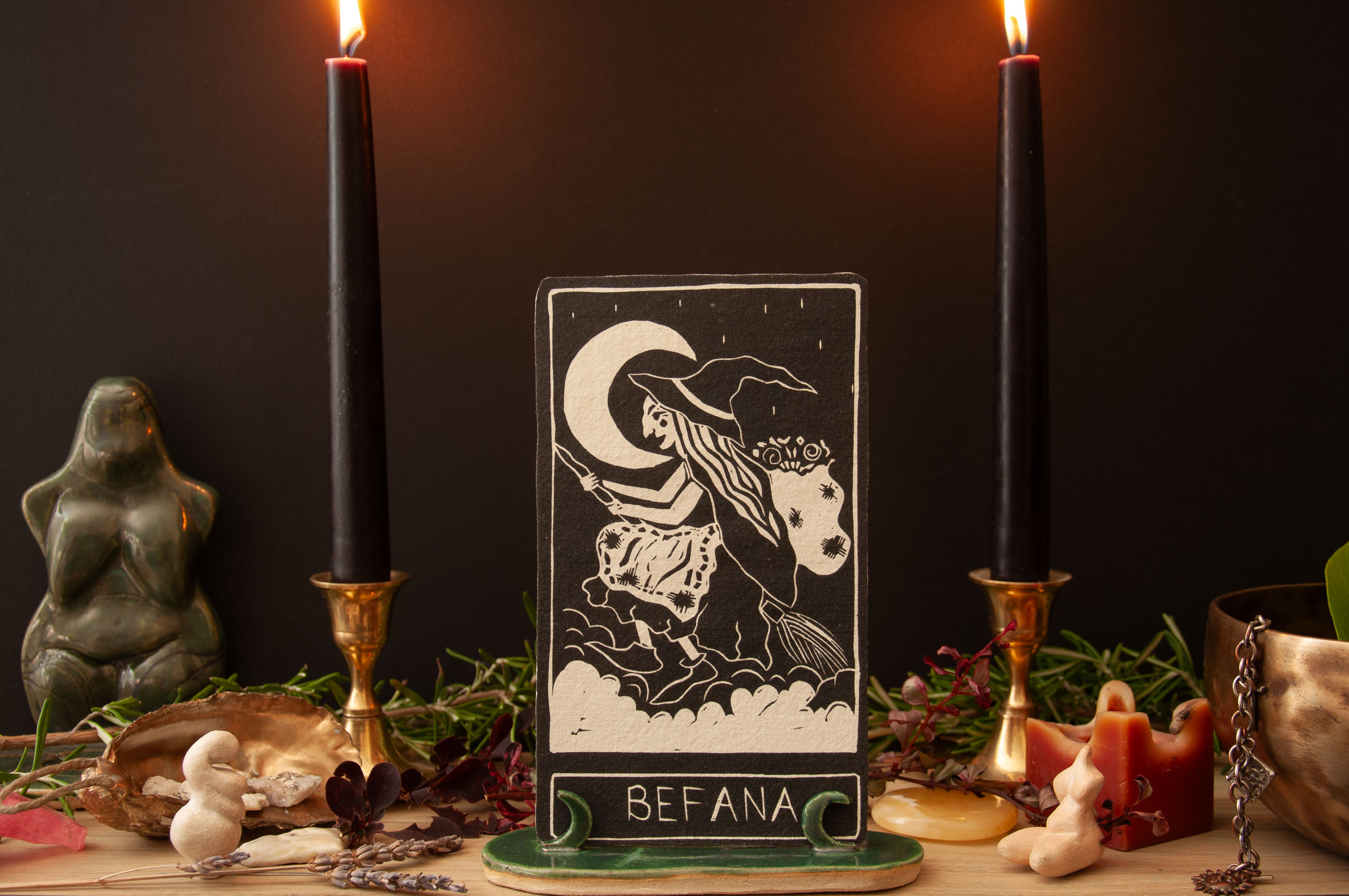 befana altar A linocut oracle card on a green moon stadn with black candles and altar objects displayed around it